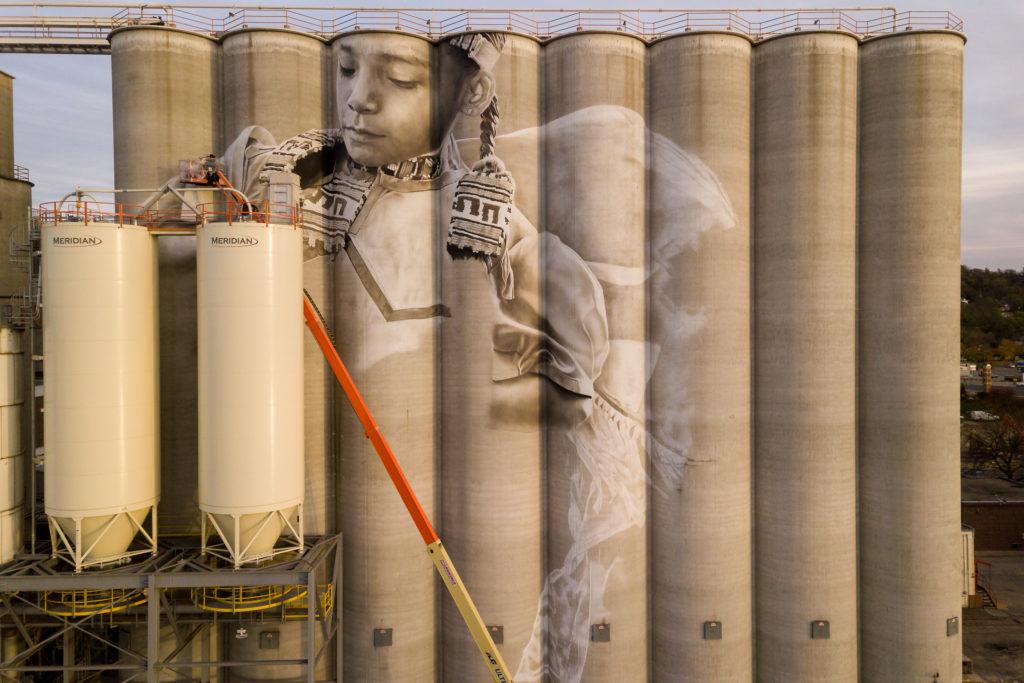 Photo by Rick Pepper - Aerial view of the in-progress Silo Art Project by Australian artist, Guido van Helten on the Ardent Mill grain silos at the gateway of Old Town, Mankato. The artist can be seen at work.