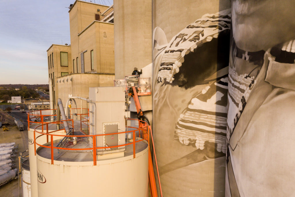 Photo by Rick Pepper - Aerial view of the in-progress Silo Art Project by Australian artist, Guido van Helten on the Ardent Mill grain silos at the gateway of Old Town, Mankato. The artist can be seen at work.