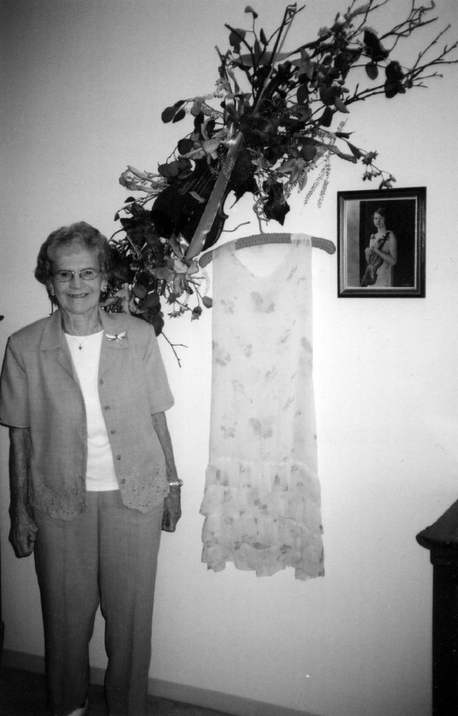 Photo by Julie Schrader - Muriel Kuebler Berndt in 2004, standing next to her violin (which was made into a floral arrangement for her wall), one of her original dance dresses and an old photograph of herself with the violin.