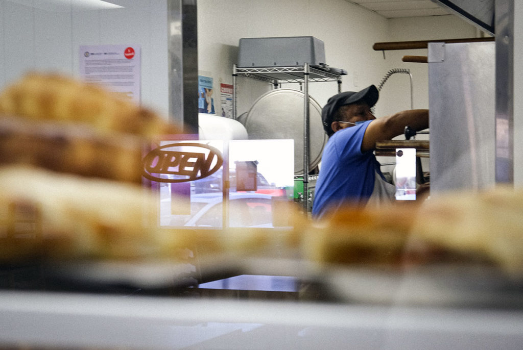 Photo by Casey Ek - Hensta places food in an oven while an ‘open’ sign reflects on a display case in Great New York Pizza.