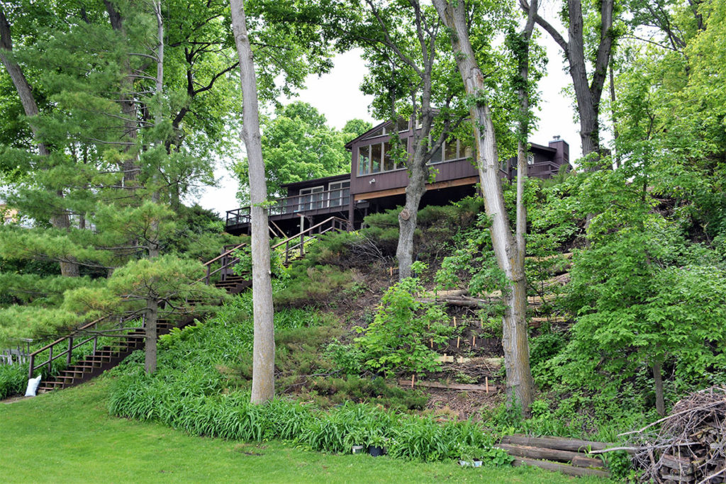 Photo by Karen J Wright - Karen's house, showing the steep slope where water can rush down into the lake. Karen has planted the hillside with native plants to help slow the water and erosion, and then added native pollinator plantings on the lakeshore to slow and absorb pollutants before going into the lake.