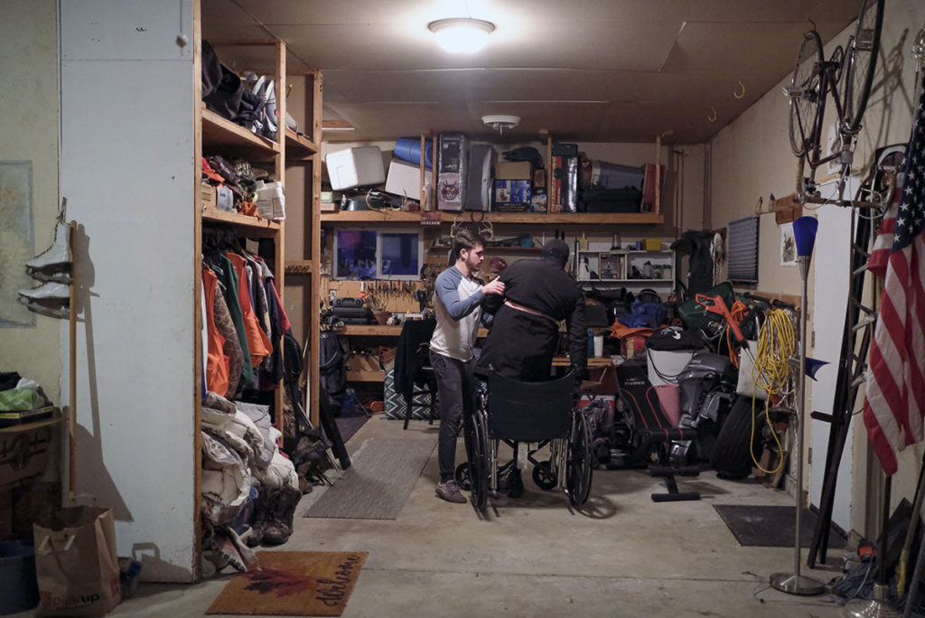 Photo by Casey Ek - Personal trainer and close friend of Chudier Dang, Alex Murphy, assists Dang out of his wheelchair in Murphy’s garage in North Mankato. Murphy said that while he is not a physical therapist, his routines for Dang come from extensive research and with the blessing of Dang’s therapist. Responsibly repeating exercises, Murphy said, is crucial in Dang’s recovery as he attempts to rebuild muscle control and neural pathways to the left side of his body.