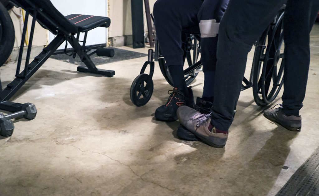 Photo by Casey Ek - Murphy braces Dang's foot as he stands up from his wheelchair. Dang’s stroke caused him to lose control of his left extremities, meaning simple activities such as standing, or walking could result in severe hyperextensions in his legs.